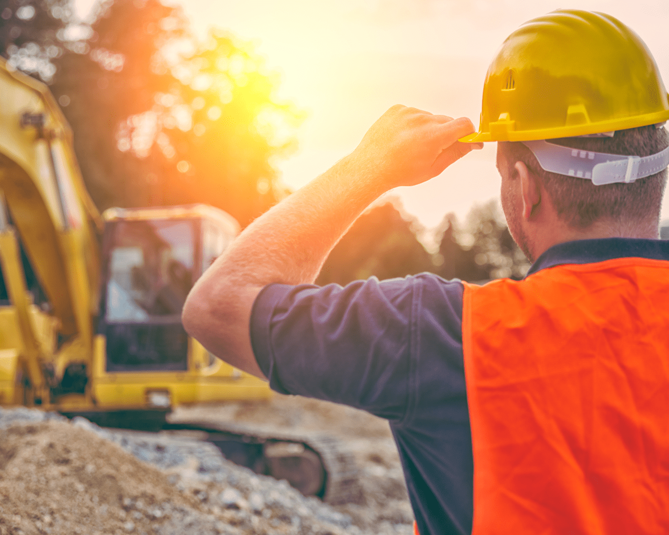 Shot from behind of construction worker with protective gear on, watching man working in excavator as the sun rises.