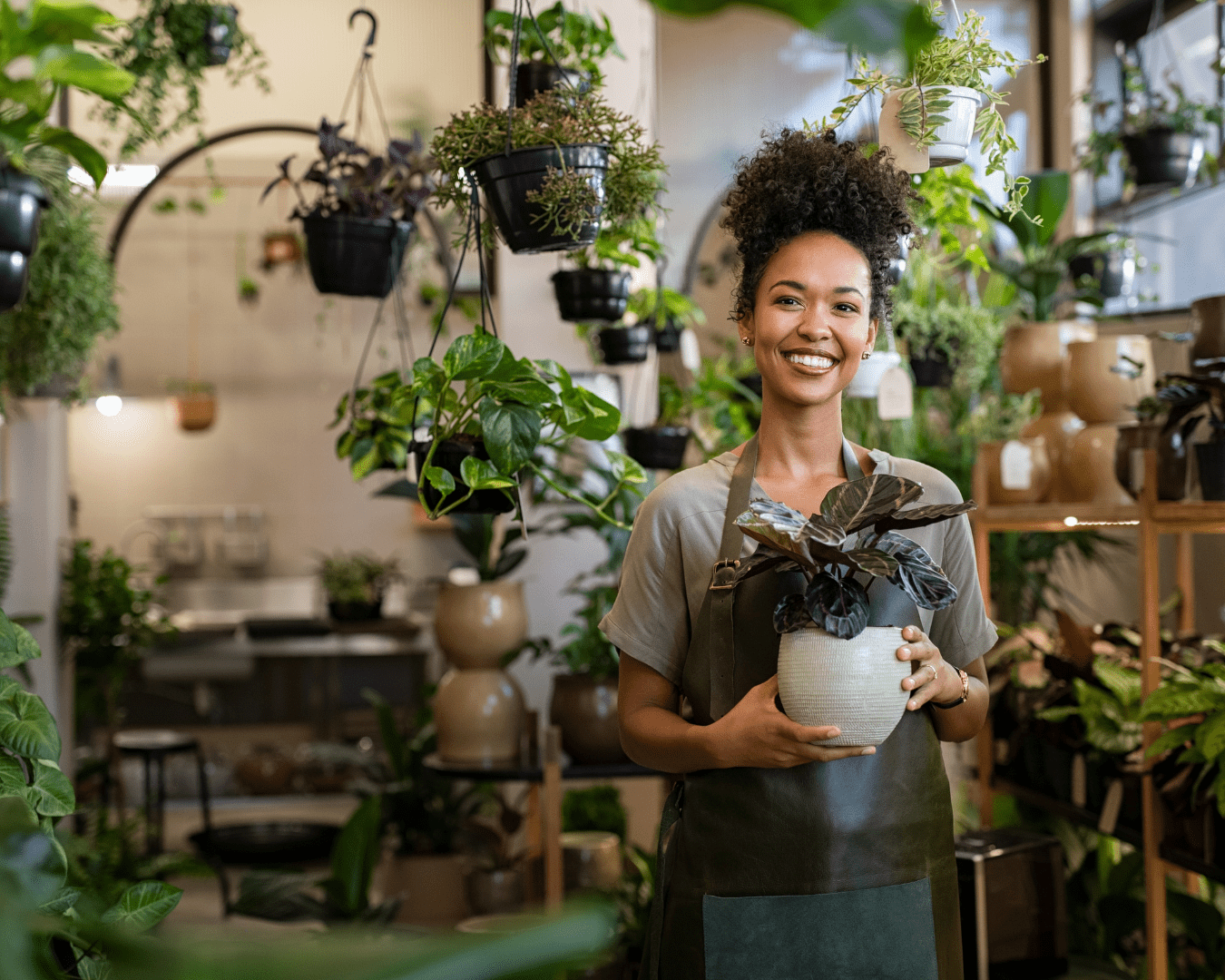 Young adult female business owner holding plant in her garden center surrounded by various plants. She's holding plant and wearing apron, smiling at camera.