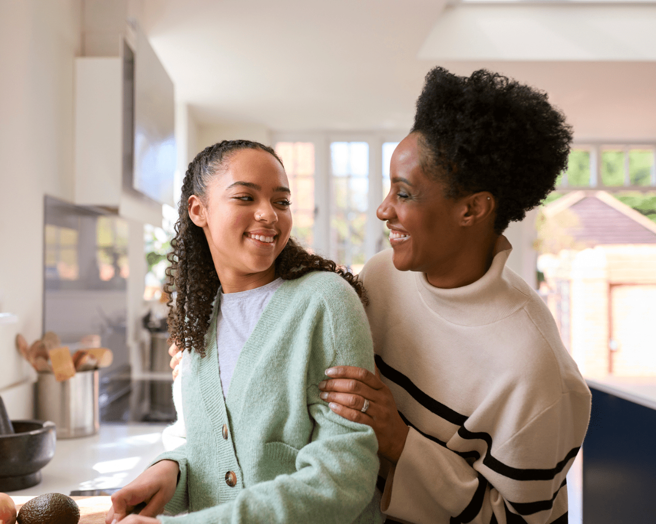 Young adult black female in kitchen cooking with her mother behind her smiling at her with her hand on her arm.